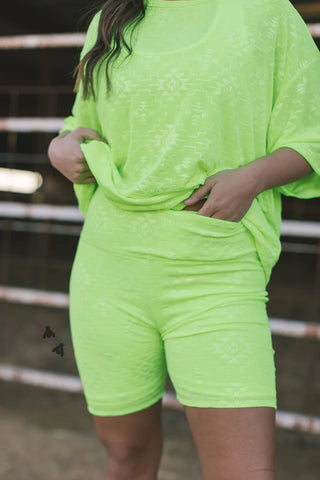 A person is wearing a neon green patterned outfit from the Western Edge Boutique's Chillville Collection, featuring a loose T-shirt and Chillville Highlighter Biker Shorts made from comfy stretch material. They stand in front of a metal fence, adjusting the shirt with their left hand.