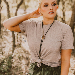 A woman stands outdoors in a sunlit forest, wearing the Brendalyn Basic Tan Top from 2 Fly, which complements her green pants perfectly. She has short blonde hair and accessorizes with hoop earrings and a long necklace. With a contemplative expression on her face, she gazes to the side while one hand lightly touches her neck.