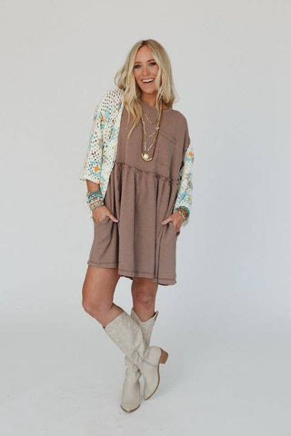 A woman with long blonde hair stands against a plain backdrop, smiling. She wears a Meadow Subtle Escape Textured Dress by Three Bird Nest, which features colorful, patterned long sleeves. She completes the look with white boots and layered necklaces. Her hands rest in the pockets of her dress, exuding effortless boho western vibes.
