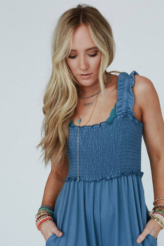 A woman with long blonde hair is wearing the Three Bird Nest's "Nothing Compares Smocked Ruffle Jumpsuit" in Denim Blue, which features wide pant legs and ruffle straps. Crafted from lightweight stretch fabric, she complements the look with layered necklaces and multiple bracelets on both wrists. She appears to be looking down, with her hands in the jumpsuit pockets against a plain white background.
