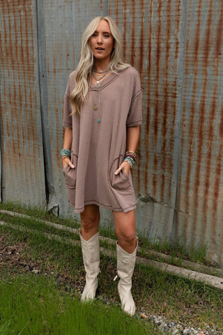 A woman with long blonde hair is standing on grass in front of a rusted corrugated metal wall. She is wearing the Brooklyn Soft Touch Tunic Dress by Three Bird Nest, which features front pockets, multiple necklaces and bracelets, and white knee-high boots. She has her hands in her pockets.