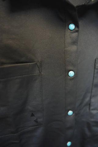 Close-up image of the LEATHER LUX * MIDNIGHT button-down shirt by 2 Fly, featuring TURQ PEARL SNAPS. The black shirt has two partially visible front pockets with a small, faint black butterfly design printed near the bottom of one pocket. The fabric appears smooth and slightly reflective.