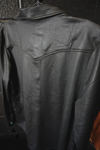 A black LEATHER LUX * MIDNIGHT jacket from 2 Fly hangs on a hanger. The image showcases the back of the jacket, emphasizing its smooth texture and stitching details on the shoulders and upper back. Crafted from luxurious leather, it features intricate TURQ PEARL SNAPS. The background is dark and out of focus.