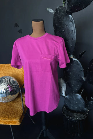The HANDY AS A POCKET* POP T-shirt from Western Edge Boutique, a vibrant pink closet staple, is showcased on a mannequin against a dark backdrop. Nearby, a tall cactus plant introduces texture to the scene. A reflective disco ball sits on a mustard yellow chair, seamlessly merging modern and western retro styles.