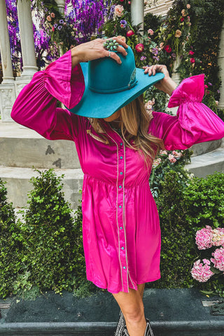 A woman in a vibrant pink BONANZA BUTTON SATIN BUTTON DOWN DRESS by 2 Fly, featuring puffy sleeves, is standing in a garden setting adorned with green plants and colorful flowers. She is wearing a teal wide-brimmed hat, partially covering her face, and holding it with both hands.