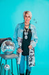 A person with short blonde hair stands against a bright blue background, wearing 2 Fly's Mackey Western Chambray and Cow Print Dress layered over a black shirt and paired with chambray denim jeans. They accessorize with a chunky necklace, holding the dress's lapels while a disco ball perches on a stool next to them.