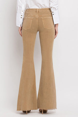 The image shows the back view of a person wearing VERVET by Flying Monkey's Ellis High-Rise Super Flare Jeans in stretch denim with frayed hems. The flared brown pants feature two back pockets and are paired with a white button-down shirt. The person is standing facing away from the camera.