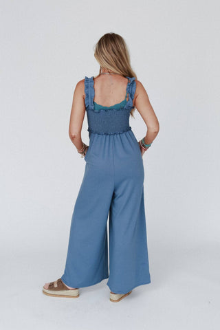 An individual stands with their back to the camera, highlighting the Nothing Compares Smocked Ruffle Jumpsuit - Denim Blue by Three Bird Nest. The sleeveless jumpsuit, made from lightweight stretch fabric, features wide-legged pants and ruffled straps. The person accessorizes with bracelets on each wrist and brown platform sandals, against a plain white background.