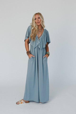 A woman with long blonde hair poses in the Electric Blue On Repeat Open Back Ruffle Jumpsuit by Three Bird Nest. The light blue, flowy jumpsuit with short sleeves features a relaxed and loose silhouette that fits her perfectly as she places her hands in its pockets, accessorized with multiple bracelets. She completes the look with patterned slide sandals against a plain white background.