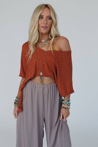 A woman with long blonde hair stands against a plain background. She is wearing a relaxed boho Kingsley Warm Nights V Neck Top in Cayenne by Three Bird Nest, featuring an off-the-shoulder style, and grey wide-leg pants. Multiple beaded bracelets and necklaces adorn her, while she gazes slightly to the side with a neutral expression.