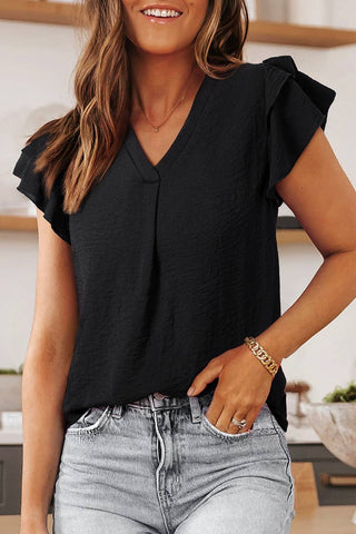 A woman with long brown hair, smiling and wearing the Pepper Women Casual Solid V Neck Ruffle Short Sleeve Blouse by Aapparella in black, along with light gray jeans, a gold bracelet, and a necklace. She is standing in a modern, light-colored kitchen with shelves and plants in the background.