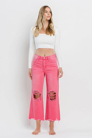 A woman with long blonde hair stands barefoot against a plain white background. She is wearing a white long-sleeve crop top and the Rosie 90's Super High Rise Crop Flare Hot Pink Jeans by VERVET by FLYING MONKEY, featuring distressed rips at the knees and raw hems, reminiscent of 90's vintage jeans.