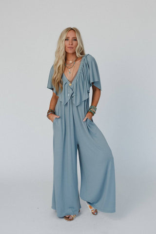 A woman with long blonde hair stands against a plain white background wearing an Electric Blue On Repeat Open Back Ruffle Jumpsuit by Three Bird Nest, made of lightweight stretch knit fabric. She accessorizes with multiple necklaces and bracelets, her hands casually in her pockets, and she completes the look with open-toe shoes.