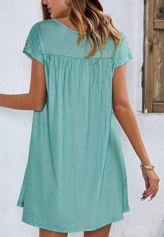 A woman with light brown hair stands facing away, wearing a loose, multicolor Sonnet Floral Boho Print Dress from Anna Kaci Wholesale. The dress features a gathered yoke and falls above her knees. She is accessorized with gold bracelets and hoop earrings. The background includes a white wall and a wooden window.