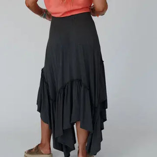 A person is shown from the back, wearing the Logan Forever Beauty Maxi Skirt in Pale Sage by Three Bird Nest, a flowing asymmetrical skirt with ruffled layers that vary in length, akin to a boho maxi skirt. It is paired with a coral-colored top. The lightweight stretch knit fabric drapes beautifully. The person is also wearing light brown shoes and bracelets against a plain white background.