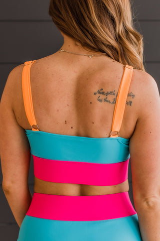 A woman is shown from the back wearing the vibrant Catching Waves Bikini Swim Top in blue, bright pink, and orange from MACK & MAI. The bikini swim top features adjustable straps for a perfect fit. She has long, wavy hair and a tattoo on her upper back. The background is a plain dark wall.