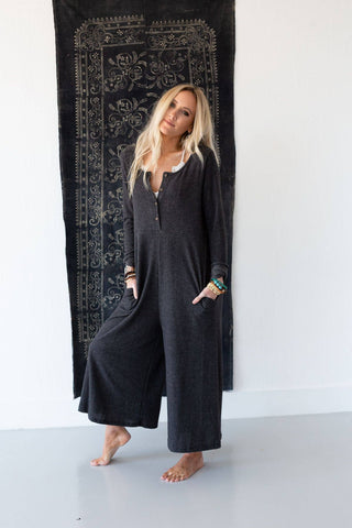 A person with long blonde hair stands barefoot in front of a tapestry, wearing the Simply Comfort Jumpsuit by Three Bird Nest. The dark gray, trendy jumpsuit is loose-fitting and features pockets. The person has one hand in a pocket and the other resting on their leg, accessorized with bracelets. The minimalistic background highlights the retro style of their outfit.