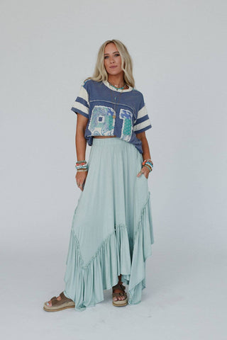A woman stands posing against a plain background. She wears a blue shirt with white stripes on the sleeves and a large "ALOHA" text on the front, paired with the Logan Forever Beauty Maxi Skirt in pale sage from Three Bird Nest, sandals, and multiple bracelets. Her long blonde hair is loose.