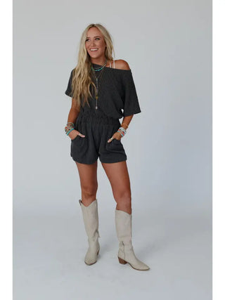 A woman with long, wavy blonde hair stands smiling. She wears an off-the-shoulder Covey Romper in Camel by Three Bird Nest, featuring an elastic waistband, beige cowboy boots, and multiple turquoise and beaded accessories. She has her hands in the pockets of her romper against a plain white background.