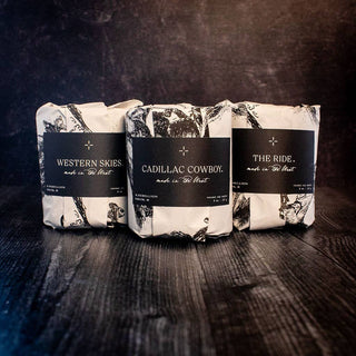 Three blacksmith-themed soap bars wrapped in rustic paper with black labels lay on a dark wooden surface, evoking the charm of The Faith Candle by R. Rebellion. The labels read: "WESTERN SKIES," "CADILLAC COWBOY," and "THE RIDE." A textured, shadowy background enhances the scene's allure.