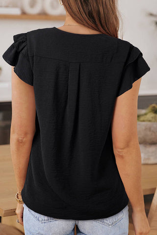 A person with long brown hair is seen from behind, wearing an Aapparella Pepper Women Casual Solid V Neck Ruffle Short Sleeve Blouse made from 100% polyester. The blouse is perfect for daily wear. They are standing indoors, in front of a wooden table with plants and shelves in the background.