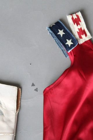 Close-up of THE BETSY ROSS by 2FLY, a red garment with embroidered straps showcasing American flag patterns. One strap features blue fabric adorned with white stars, while the other displays red zigzag patterns on a white background. The satin stretch body is partially visible on the left against a gray background.