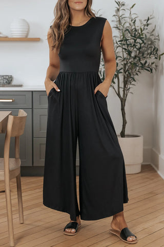 Decked out in the Quincey Black Open Back Wide Leg Jumpsuit from Western Edge Boutique, a person stands indoors with hands tucked into pockets. The sleek, sleeveless design of the breathable fabric perfectly complements the kitchen backdrop, which includes a chair and a potted plant.