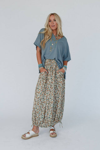 A woman with long blonde hair stands against a plain background. She is wearing a loose-fitting blue top, the Solstice Song Cinched Skirt - Khaki by Three Bird Nest featuring floral prints, layered necklaces, multiple bracelets, and white sandals. Her hands are in her skirt pockets, and she poses confidently.
