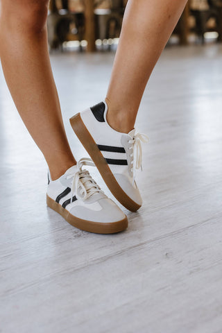 A person wearing Ness Striped Lace Up Flat Sneakers by Liam & Company stands on a light wooden floor. The sneakers have black stripes, gum soles, and are tightly laced. The person's legs are slightly bent.