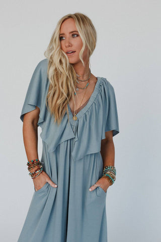 A woman with long, wavy blonde hair wears the Electric Blue On Repeat Open Back Ruffle Jumpsuit by Three Bird Nest. She has layered necklaces and beaded bracelets on both wrists. With her hands in her pockets, she gazes off to the side against a plain white background.