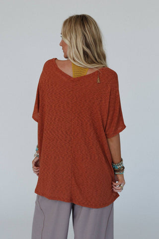 A person with long blonde hair is standing facing away from the camera, wearing the oversized Kingsley Warm Nights V Neck Top in Cayenne by Three Bird Nest. The top has short sleeves and pairs perfectly with grey pants that exude a relaxed boho western style. Multiple bracelets adorn both wrists, and a yellow top or sports bra peeks out beneath the comfortable ribbed knit fabric of their ensemble.