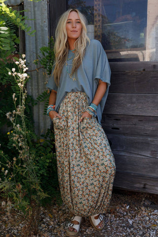 A woman with long blonde hair stands outdoors in front of a rustic wooden structure. She is wearing a loose-fitting gray top, the Solstice Song Cinched Skirt in khaki by Three Bird Nest, and white sandals. With her hands in her pockets and adorned with multiple turquoise jewelry pieces, she radiates a relaxed vibe.