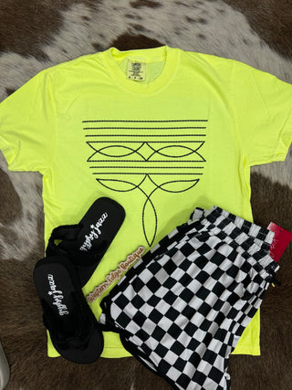 Vibrantly displayed on a brown and white cowhide rug is the Tayce Neon Boot Stitch from Western Edge Boutique, a bold t-shirt in bright yellow adorned with an abstract black line design. It pairs sleekly with black and white checkered shorts, while nearby sit elegant black slippers featuring cursive text.