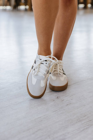 Close-up of a person wearing Liam & Company's Ness Striped Lace Up Flat Sneakers. The design features brown soles and a simple white style with laces, standing on a light-colored floor and focusing on the lower legs and feet.