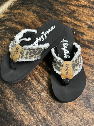 A summer essential, the Wild Leopard Flip Flops from Western Edge Boutique boast fuzzy leopard-print straps complemented by brown leather accents. The insoles feature the "Gypsy Jazz" branding in white cursive text, all elegantly set against a textured brown and black surface.
