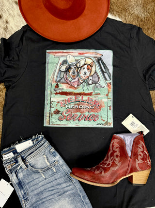 A "Heeler Truck Ride" t-shirt from Western Edge Boutique, featuring quirky prints of two dogs in a truck, paired beautifully with jeans and red cowboy boots for that authentic Western Edge style. Topped off with a red cowboy hat, the ensemble proudly displays the bold text "Heeler Herding Service" for the ultimate TROUBLE makers.