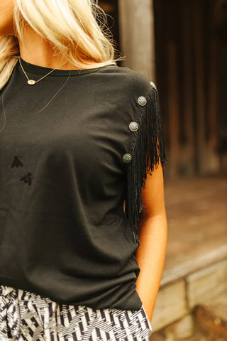 A person with long, blonde hair is wearing the "Get Fringey with it Black Top" from 2 Fly, which features fringe sleeves and button details along the shoulder seam. They are also sporting patterned pants with their hands in pockets. The background is blurred, featuring a wooden structure.