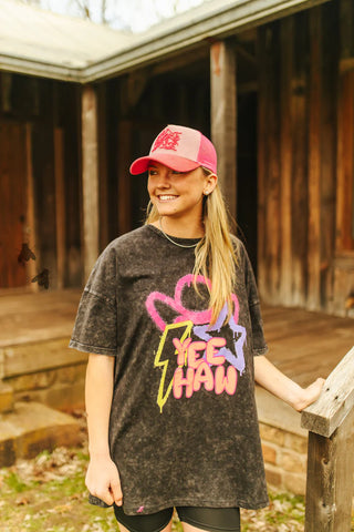 A person stands on a wooden porch, smiling and wearing the "ELECTRIC GRINGA" tee by 2 Fly that features neon colors, vibrant designs, and the text "YEE HAW" on the front. They are also sporting a pink and red cap while leaning on a wooden railing, embracing summer fun against the backdrop of a rustic wooden building.