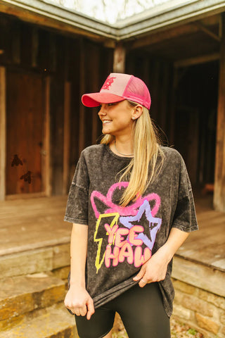 A person wearing a red baseball cap and an oversized black t-shirt featuring neon-colored "Yee Haw" text from 2 Fly's ELECTRIC GRINGA line stands outside a wooden building. With long blonde hair and a smile, they epitomize summer fun against the backdrop of a rustic porch with steps, perfect for any warm weather wardrobe.
