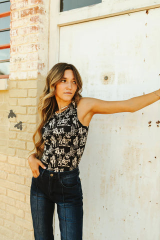A young woman with long, wavy hair strikes a pose against an old, weathered white door with a brick wall nearby. She is wearing the 2 Fly DUSK DESERT TANK and high-waisted dark jeans—an all-season wear look. Two black butterflies are flying near her in the image.