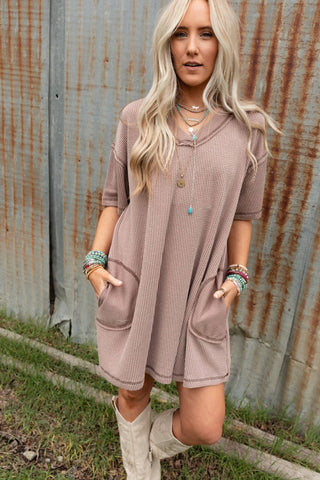 A woman with long blonde hair stands outside against a corrugated metal wall, wearing the Brooklyn Soft Touch Tunic Dress from Three Bird Nest. This boho-chic dress is made of comfortable stretch fabric and features a loose-fitting, short-sleeved design in taupe with two front pockets. She pairs the dress with multiple bracelets, layered necklaces, and knee-high white boots.