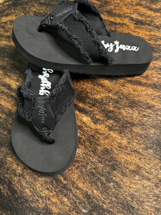 A pair of Slate Black Flip Flops featuring frayed edges on the straps, offering ultimate comfort. The sandals have the brand name "Western Edge Boutique" inscribed in white on the insole. They are placed on a surface with a brown and black pattern resembling animal fur.