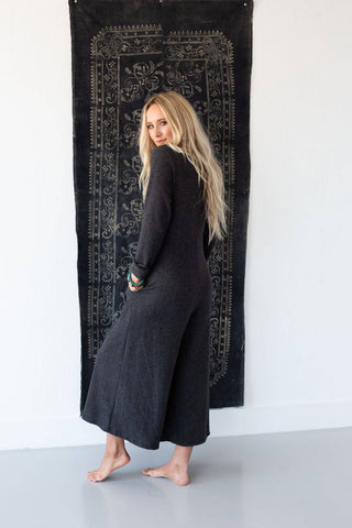 A person with long, wavy blonde hair stands barefoot on a light grey floor, clad in the trendy Simply Comfort Jumpsuit by Three Bird Nest, featuring long sleeves in dark grey. They are facing backward, their head turned slightly to the side. A black tapestry with intricate patterns adds a touch of retro style to the white wall behind them.
