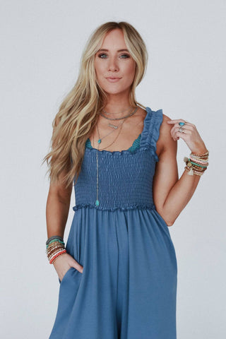 A woman with long blonde hair is dressed in the "Nothing Compares Smocked Ruffle Jumpsuit - Denim Blue" by Three Bird Nest. She is accessorized with several layered necklaces, bracelets, and rings. Standing against a plain white background, she gazes directly at the camera with one hand in her pocket and the other gently touching her hair.