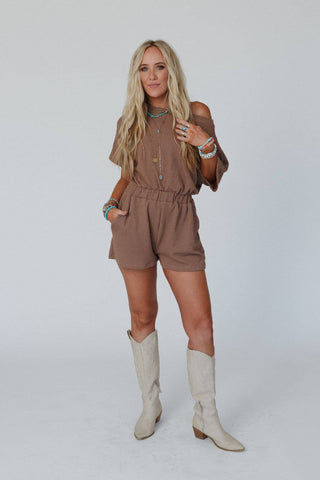 A woman with long blonde hair stands against a plain background, wearing the Covey Romper - Camel by Three Bird Nest. The light brown romper features an off-the-shoulder design, short sleeves, pockets, and an elastic waistband. She accessorizes with multiple necklaces and bracelets and wears knee-high, light-colored cowboy boots.