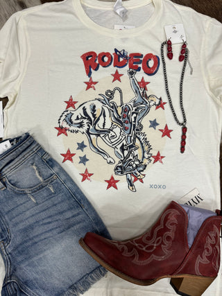 Flat lay of a white "All American Rodeo" T-shirt from Western Edge Boutique, featuring a cowboy print surrounded by denim shorts, red cowgirl boots, and matching red jewelry. The exclusive design includes stars and a bucking bronco, making it perfect for any concert ensemble. Tags are visible on all clothing items.