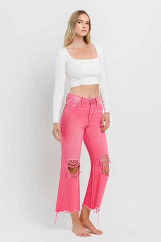 A woman with long blonde hair stands barefoot against a plain white background. She is wearing a fitted white long-sleeve crop top and Rosie 90's Super High Rise Crop Flare Hot Pink Jeans by VERVET by FLYING MONKEY, featuring ripped knees and frayed hems for that perfect 90's vintage look.