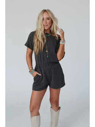 A woman with long blonde hair is wearing a dark Covey Romper - Camel by Three Bird Nest, featuring a cinched elastic waistband. She has several colorful bracelets on her wrists, a few necklaces, and is standing confidently with one hand on her hip. She's also wearing light-colored cowboy boots.