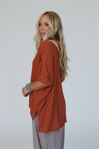 A woman with long, wavy blonde hair stands and looks over her shoulder. She wears an oversized Kingsley Warm Nights V Neck Top in cayenne from Three Bird Nest and light grey pants, exuding a relaxed boho western style. Her wrist is adorned with several colorful beaded bracelets. The background is plain and light-colored.