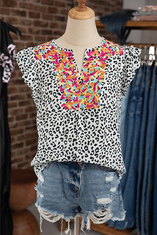 A mannequin dressed in the Aapparella Mac Women Casual Ruffle Embellished V-neck Sleeveless Top, featuring a white leopard print with colorful embroidered floral designs around the v-neckline and ruffled embellishments on the frilly sleeves. The top is paired with distressed denim shorts. The background showcases exposed brick walls and racks filled with various clothing items.